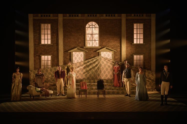Cast members dressed in flamboyant regency dress stand at various levels on stage with steps at the back leading up to the façade of a large house with a huge patterned curtain being hoisted up in front of the house.